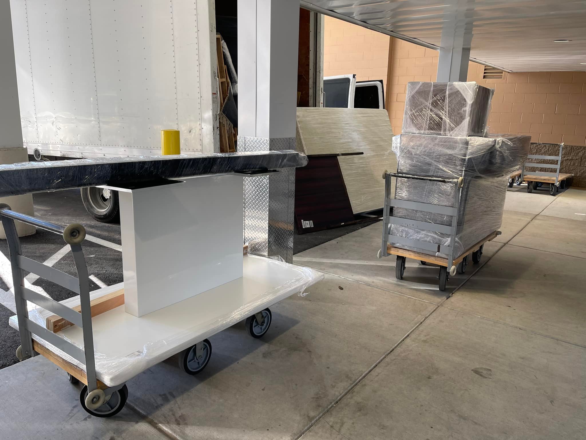 Moving dollies loaded with wrapped furniture and equipment are staged outside a building in Hayward, California, next to a parked moving truck.