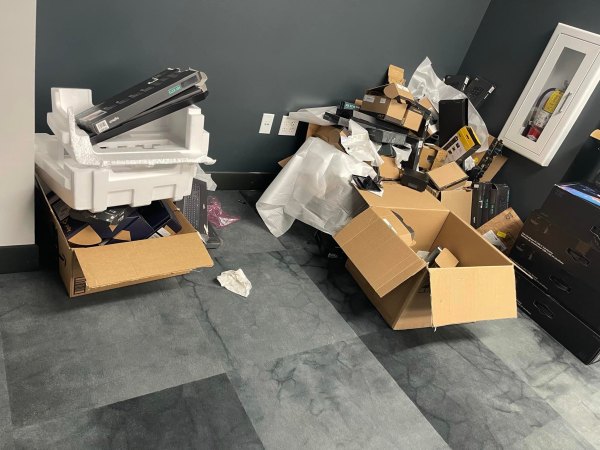A pile of discarded electronics, cardboard boxes, and packaging materials sits on a gray floor against a dark wall in an office or commercial space, awaiting junk removal.