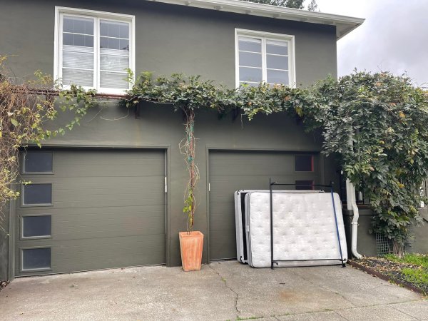 Two mattresses are propped up against a dark green garage door of a two-story house with white-framed windows.