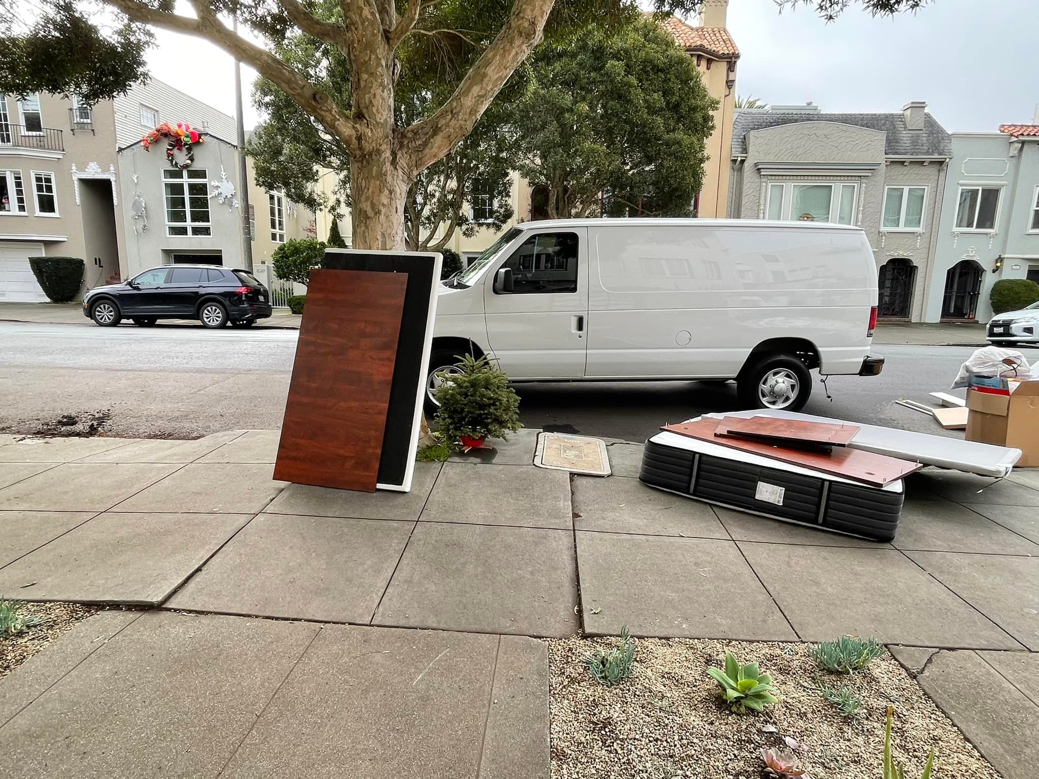 A white Ford E-Series van is parked on a residential street in Hayward, California, next to discarded furniture awaiting junk removal.
