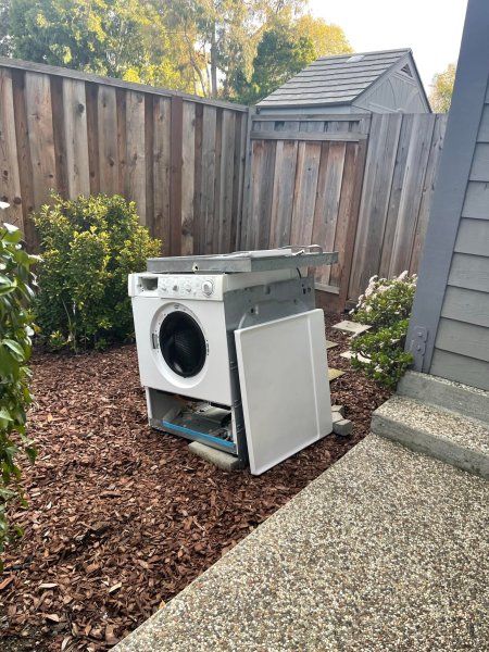 A white front-loading washing machine lies on its back on a bed of wood chips, next to a wooden fence and a small shed.