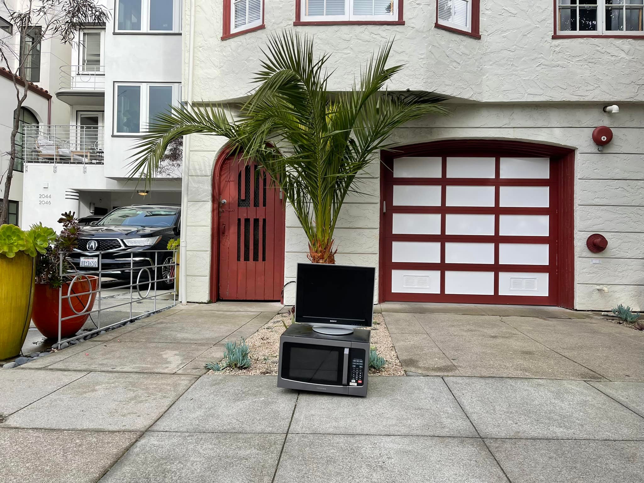 A black microwave oven and a black television set are placed on a concrete driveway in front of a white building with a red garage door.