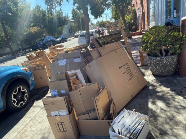 A large pile of cardboard boxes and wooden pallets is stacked on a sidewalk next to a parked blue car, awaiting junk removal on a sunny urban street.