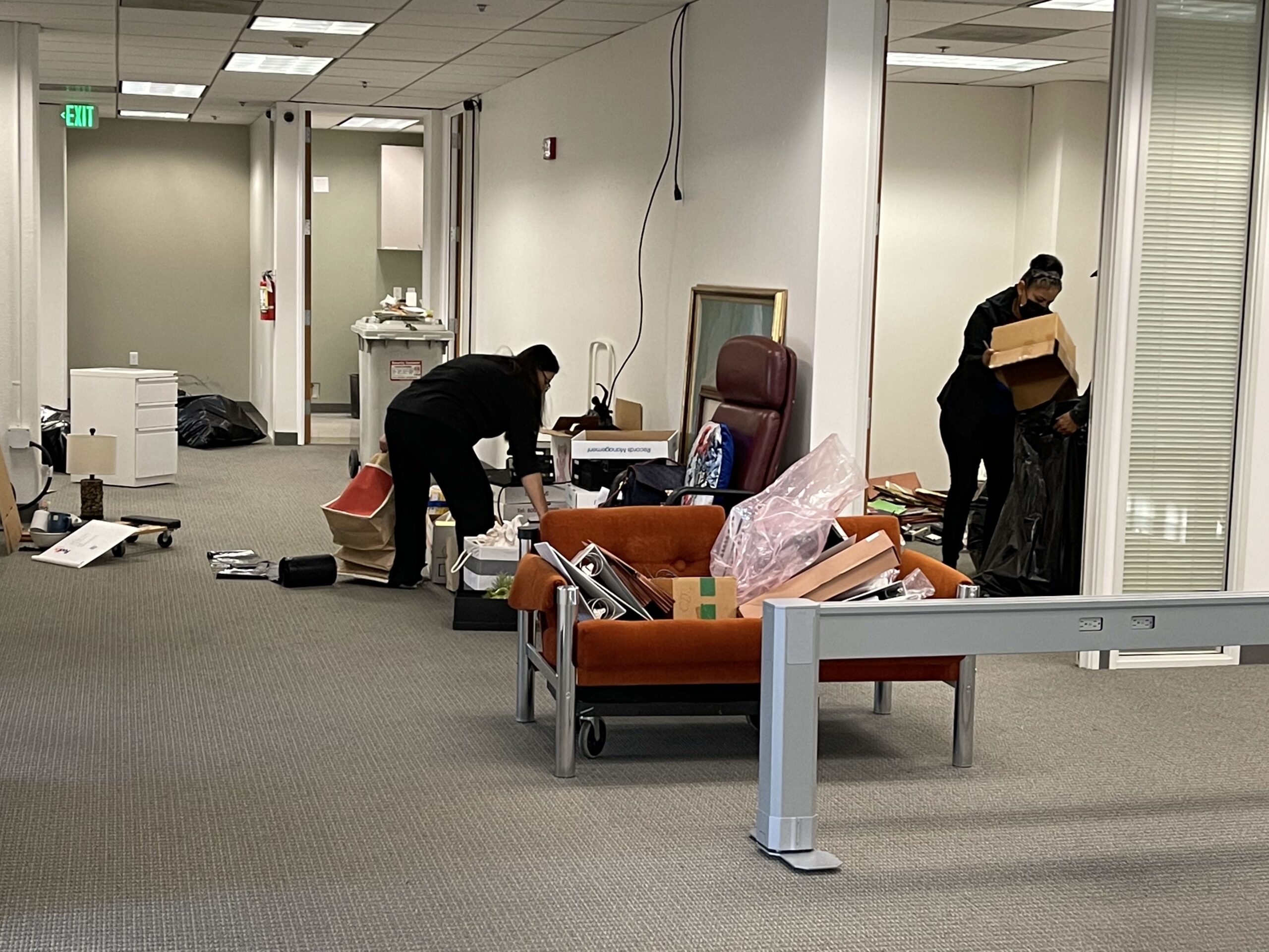 Two individuals are packing up an office space, with boxes and furniture scattered around.