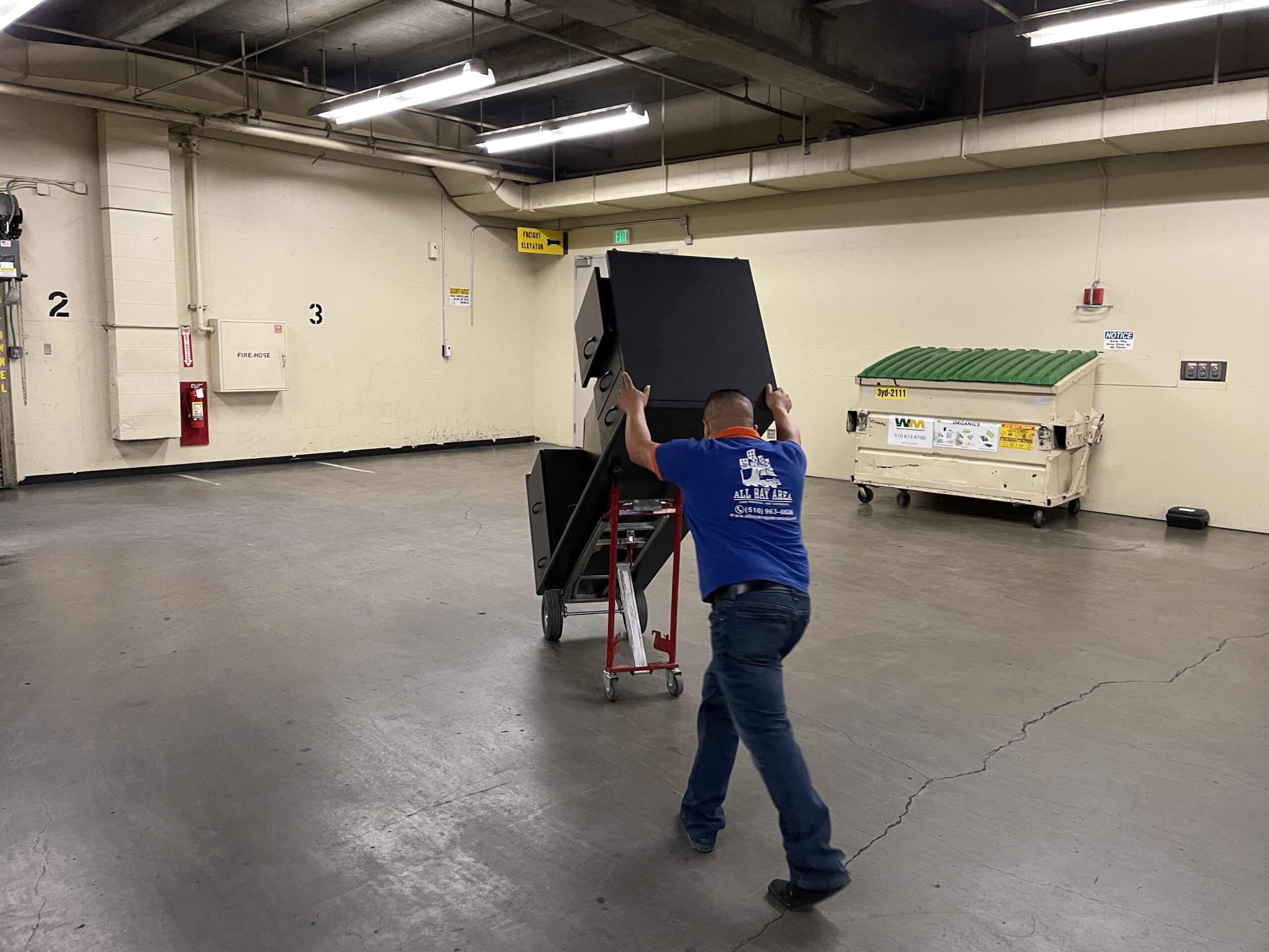 A person in a blue shirt and jeans is moving a large, black, heavy object on a wheeled platform in a warehouse or loading dock area.