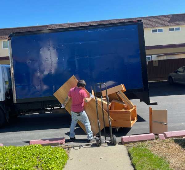A man in a red shirt and jeans is loading wooden furniture onto a blue moving truck parked on a sunny street in Hayward, California.