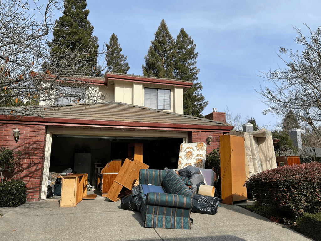 A suburban house with a two-car garage overflowing with discarded furniture and household junk spilling onto the driveway.