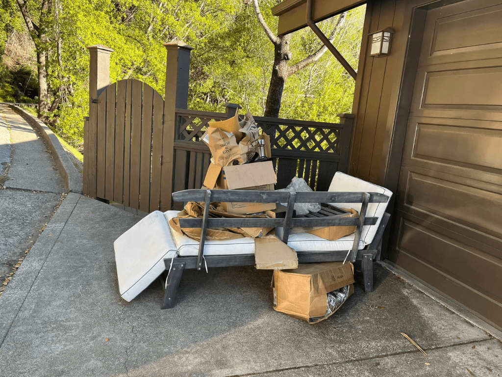 A disassembled wooden and metal patio furniture set with white cushions and several cardboard boxes piled on top, sitting on a concrete driveway.