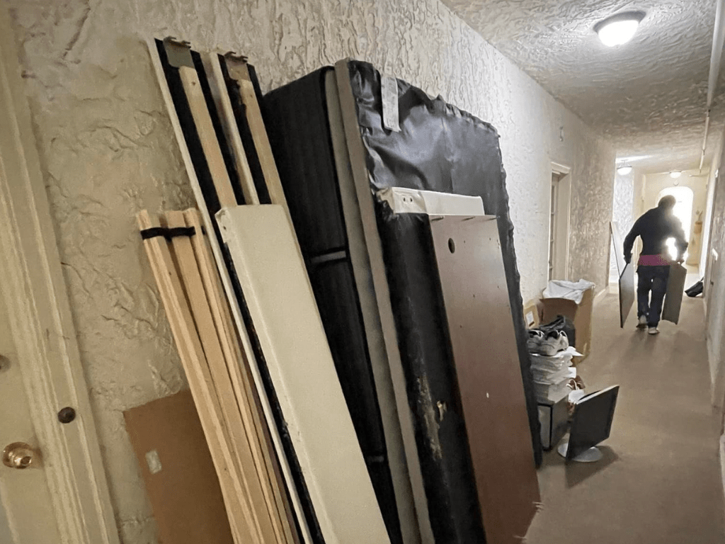 Disassembled bed frames and a mattress lean against a wall in a hallway, with a person carrying more pieces towards the front door, suggesting junk removal in progress in Hayward CA.
